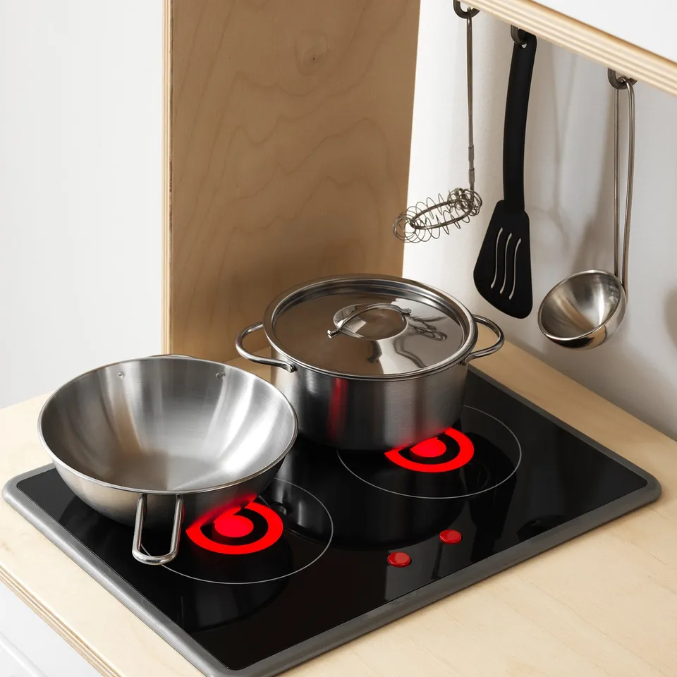 Stainless steel play kitchen utensils and pots on a toy stovetop with red burners.