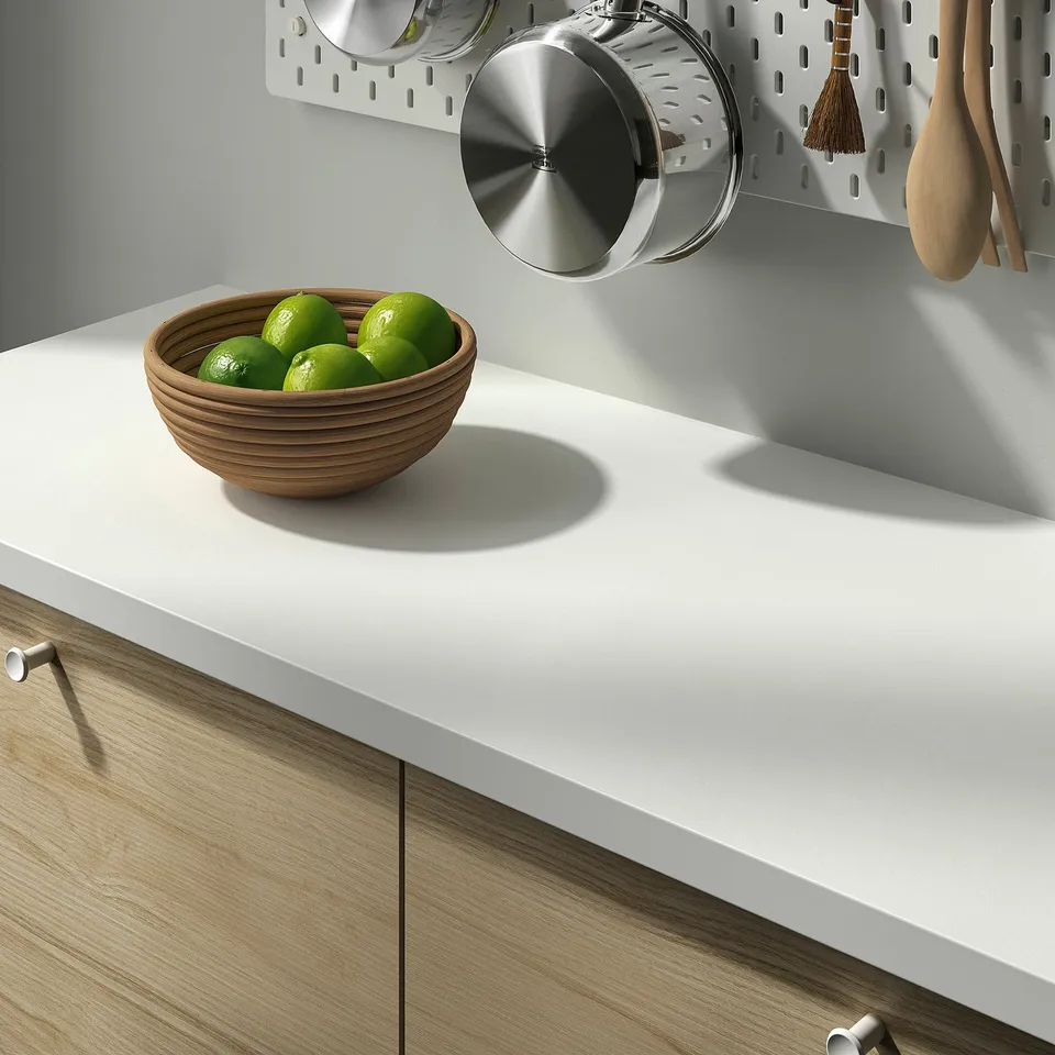 Wooden bowl filled with green limes sits on a clean, modern kitchen countertop with light wood cabinets and a stainless steel pot and utensils nearby.