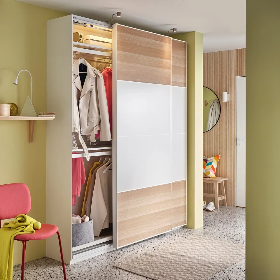 Closed wardrobe named KOMPLEMENT in beige with wooden accents, featuring sliding doors, clothes rail inside, with red chairs and yellow wall.