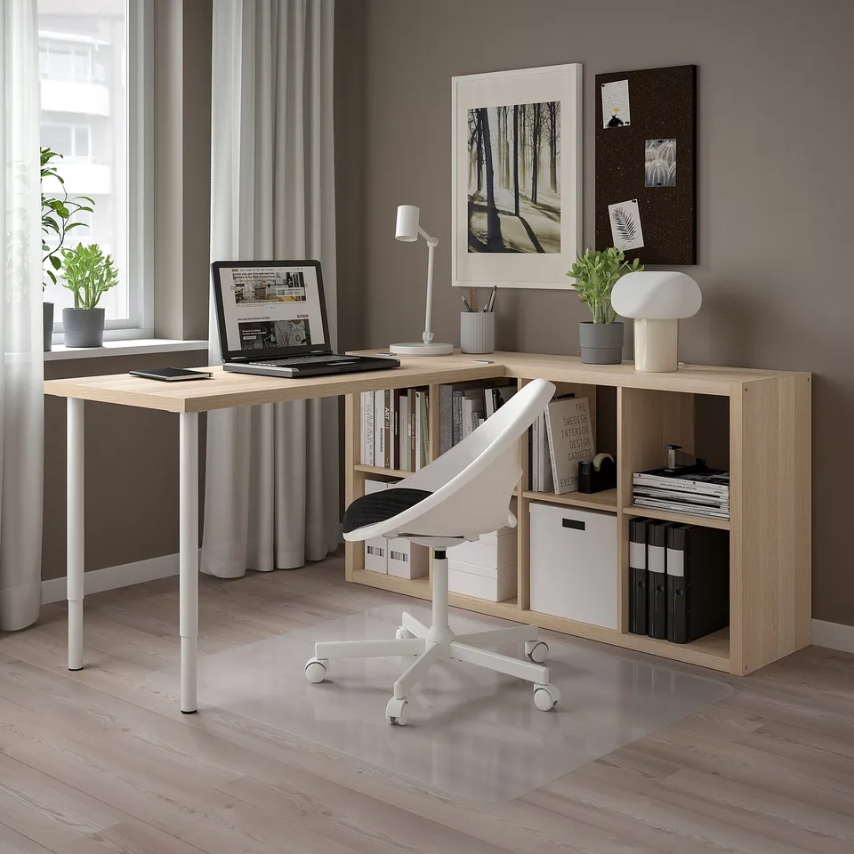Home office setup with a light wooden KALLAX desk, white chair, and shelves holding books, laptop, and plants. Grey curtains frame a large window.