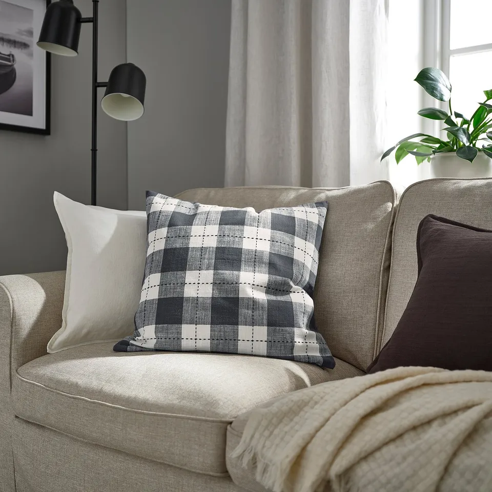 Cosy living room with beige sofa, patterned cushion, pillows, and blanket. Tall black lamp and green plant by window.