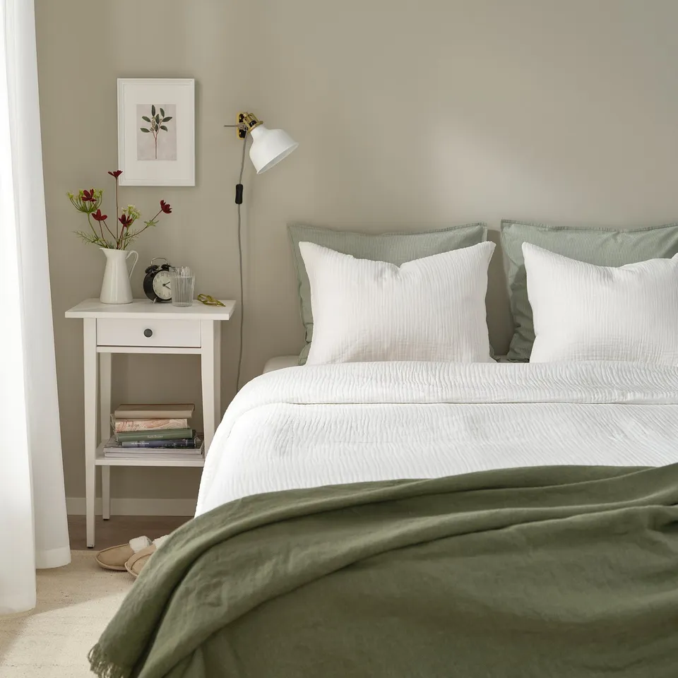 Minimalist bedroom: white bed with green blanket, bedside table, lamp, books, vase with flowers.