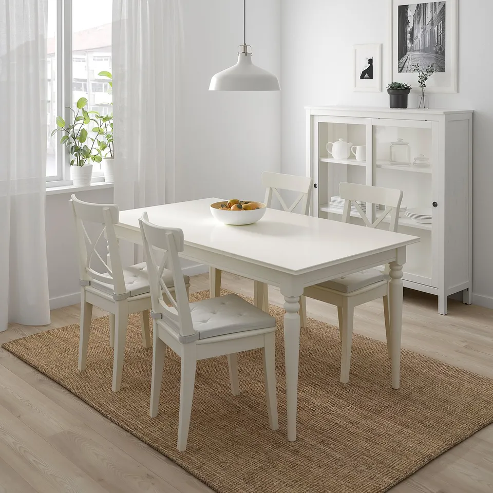 White JUSTINA dining set by window. Fruit bowl, comfort chairs, cabinet with dishes & plant.