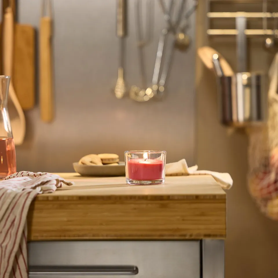 A small, red LOTSFÅGEL scented candle sits on a wooden counter, surrounded by kitchen utensils; its smaller than the nearby glass and dish.