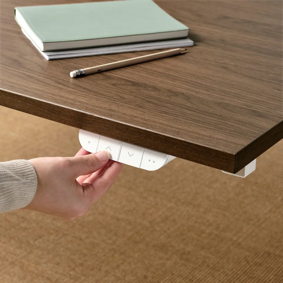 Hand presses control on MITTZON sit/stand desk. Desk is wooden with a stack of books and a pencil on top.