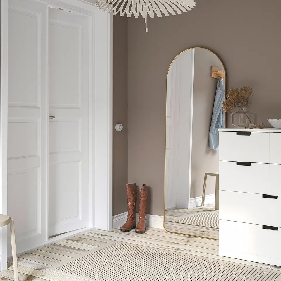 LINDBYN gold mirror with slim frame, floor to ceiling, reflecting boots on striped rug, cabinets, and door, enhancing room depth.