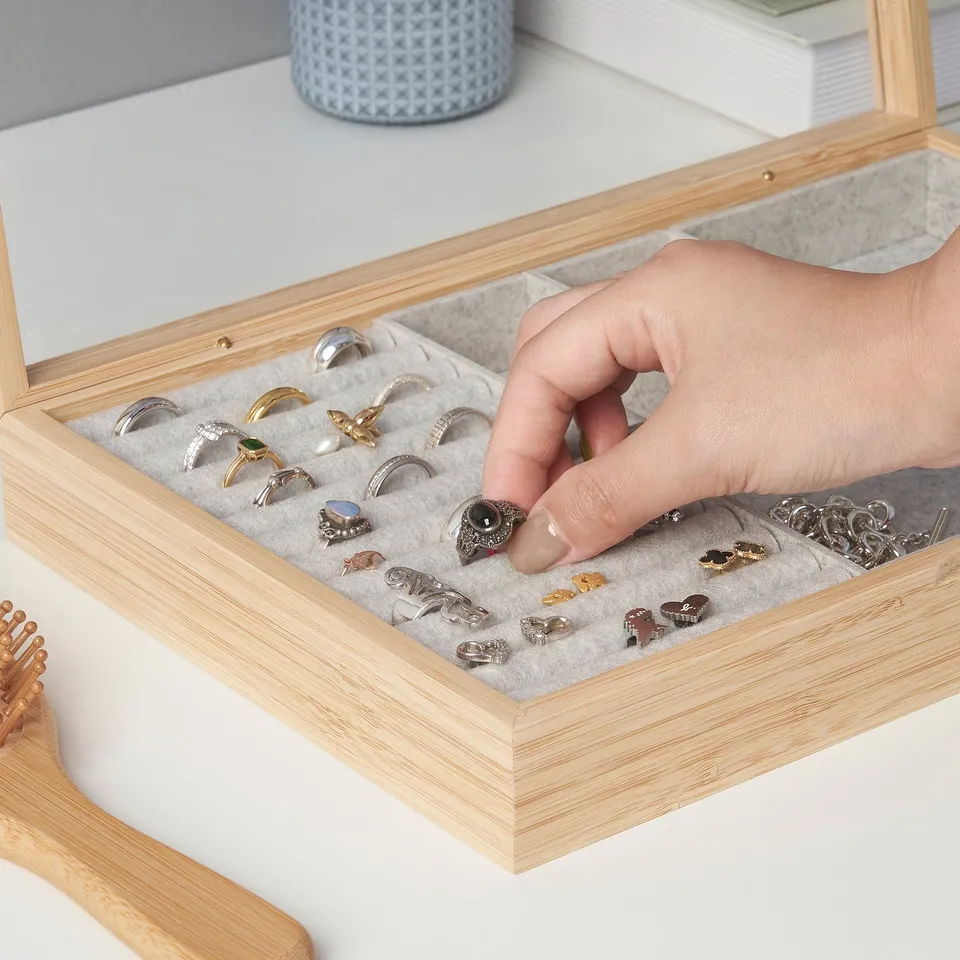 A hand selecting a ring from a wood jewellery box with compartments, displaying various rings and small accessories.