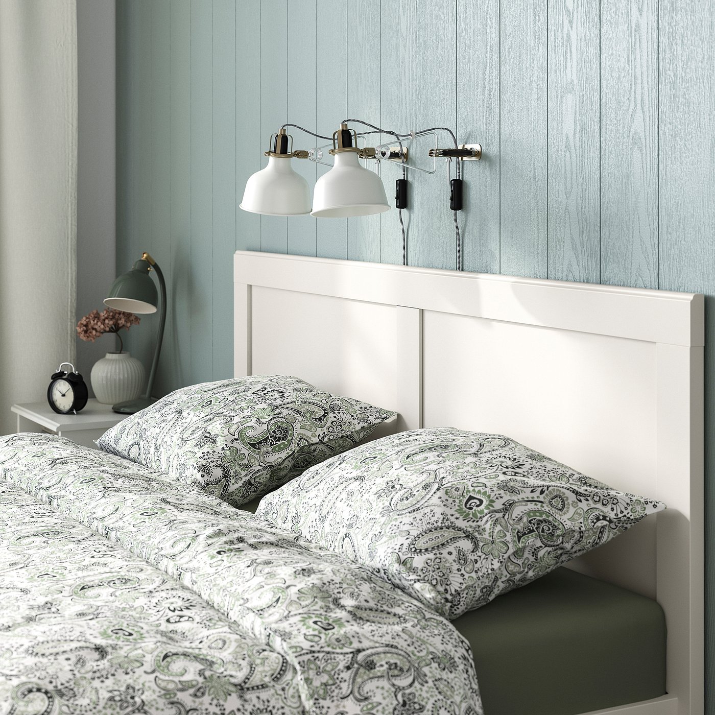 Bedroom scene: white bed with grey-green paisley bedding, adjustable wall lamps, small bedside table with clock and vase.