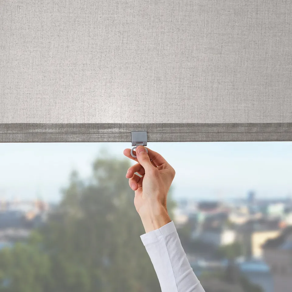 A hand lowers a cordless, sheer fabric roller blind. The blind philtres light and provides privacy. Suitable for tall windows or balcony doors.