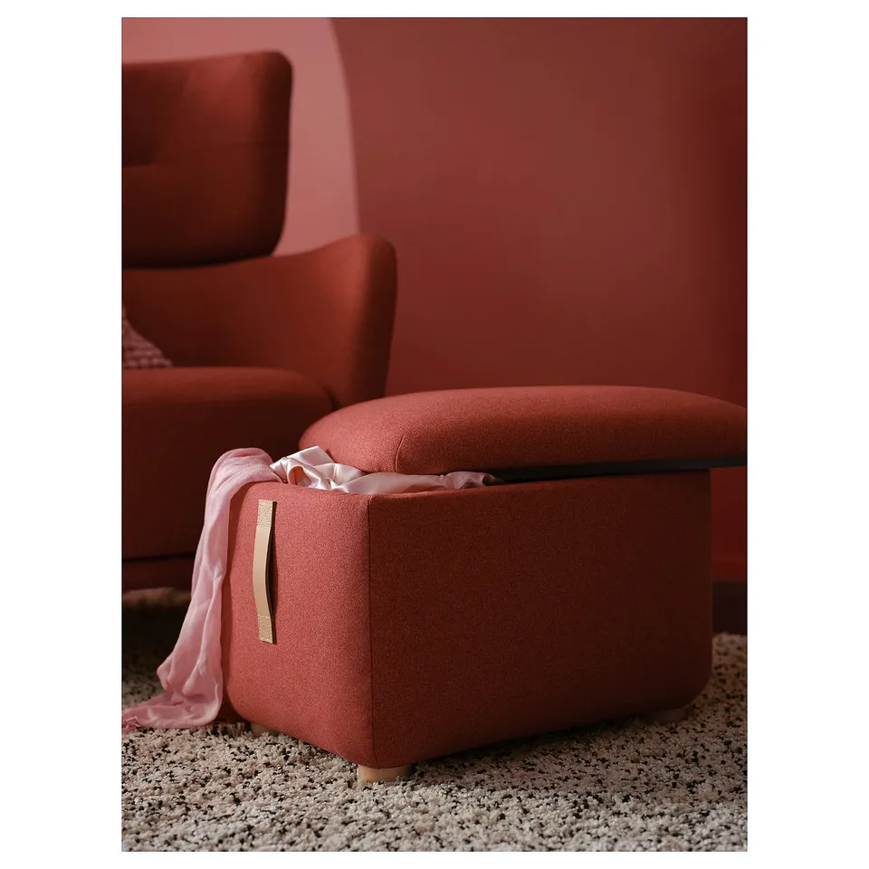 Red upholstered footstool with leather handle, placed on a textured rug, beside a matching wing chair.