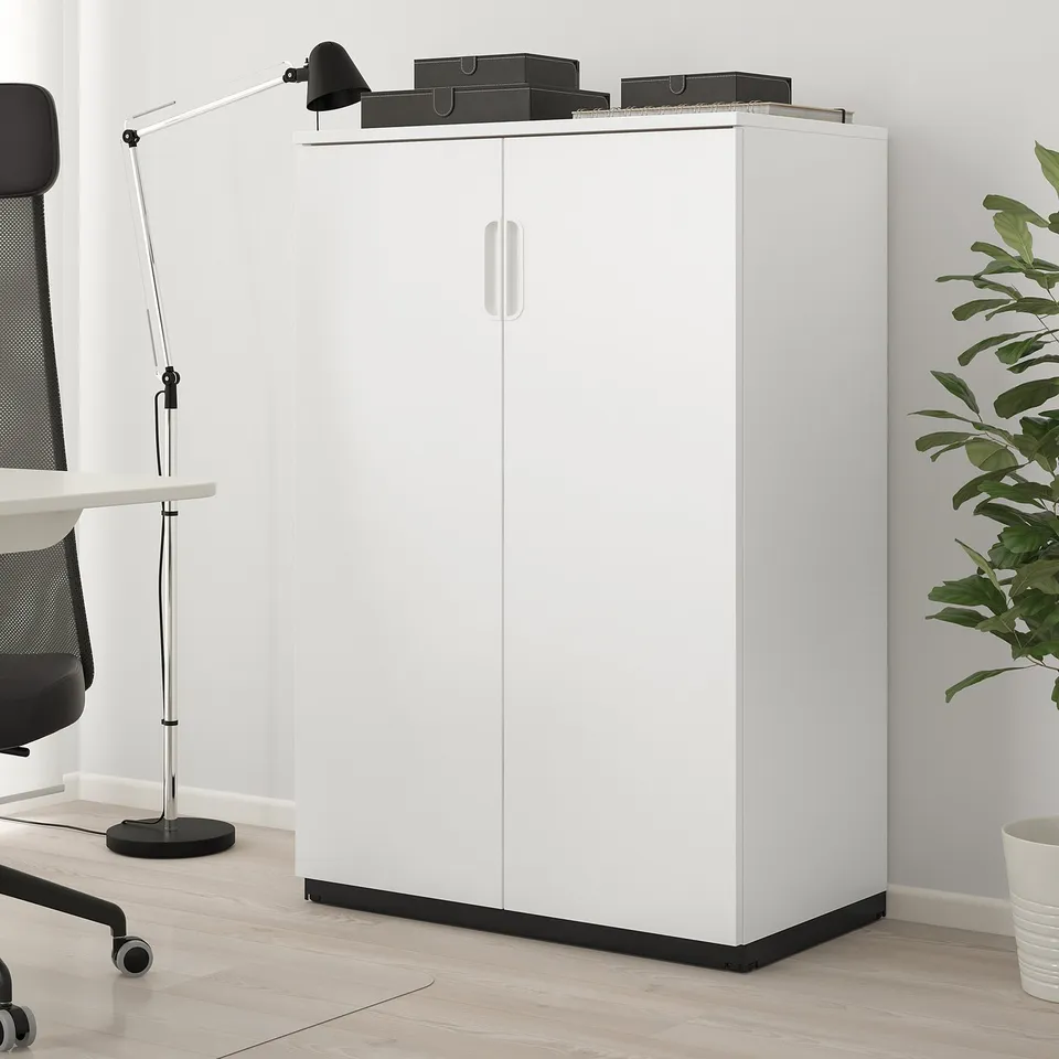 White GALANT cabinet on wood floor, tall & modern with double doors, next to black chair & desk.
