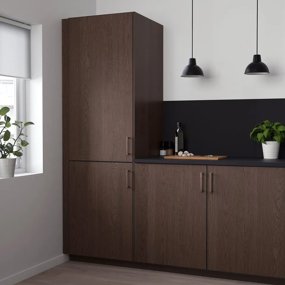 Modern kitchen with tall dark brown SINARP cabinets, sleek handles, and hanging lamps. Two potted plants and a window.