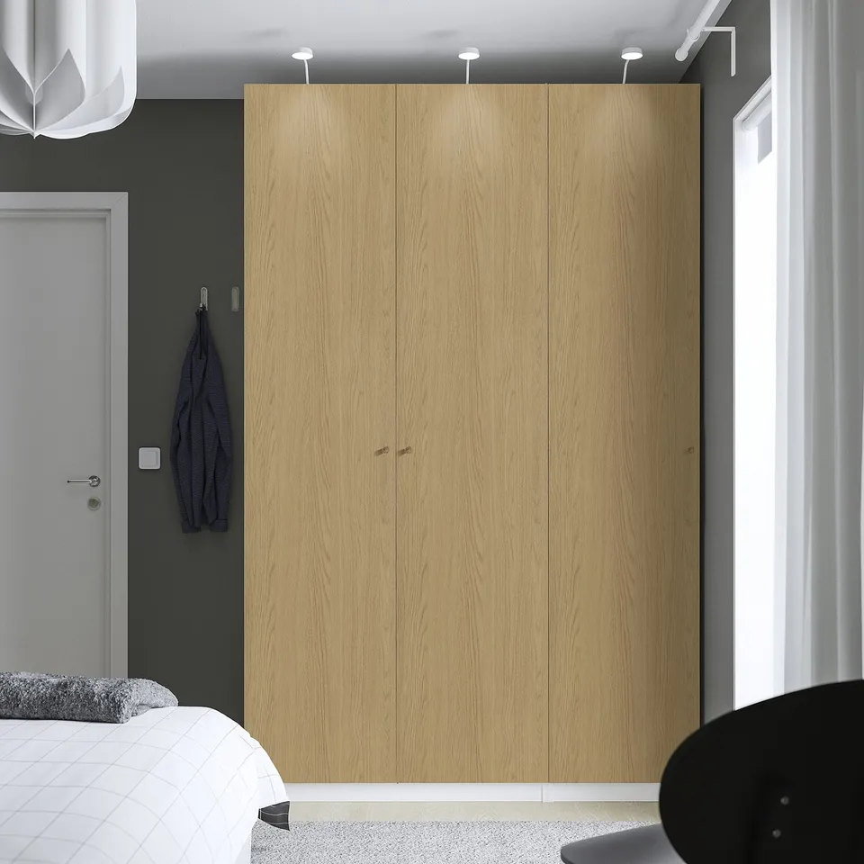 Minimalist bedroom with tall wooden PAX wardrobe, white door, blue jacket, and bed with grey pillow.