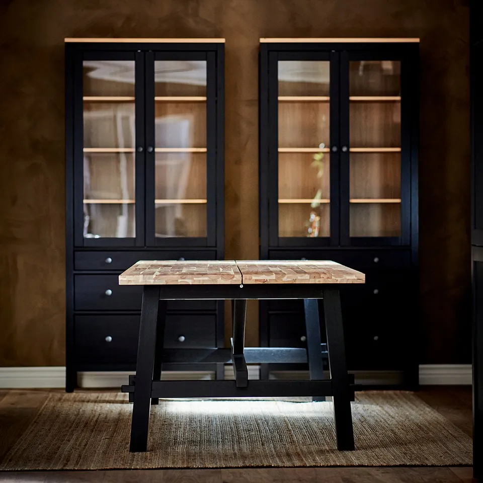 Modern dining table with black metal legs and wooden top, set against dark walls with glass-fronted cabinets.