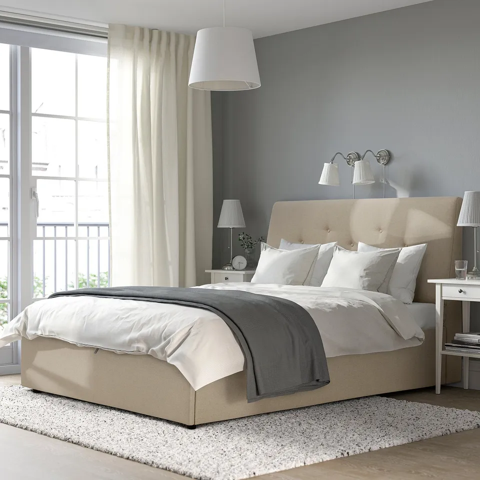 Beige IDANÄS ottoman bed, sloped headboard, white bedding, grey throw, bedside table, lamps.