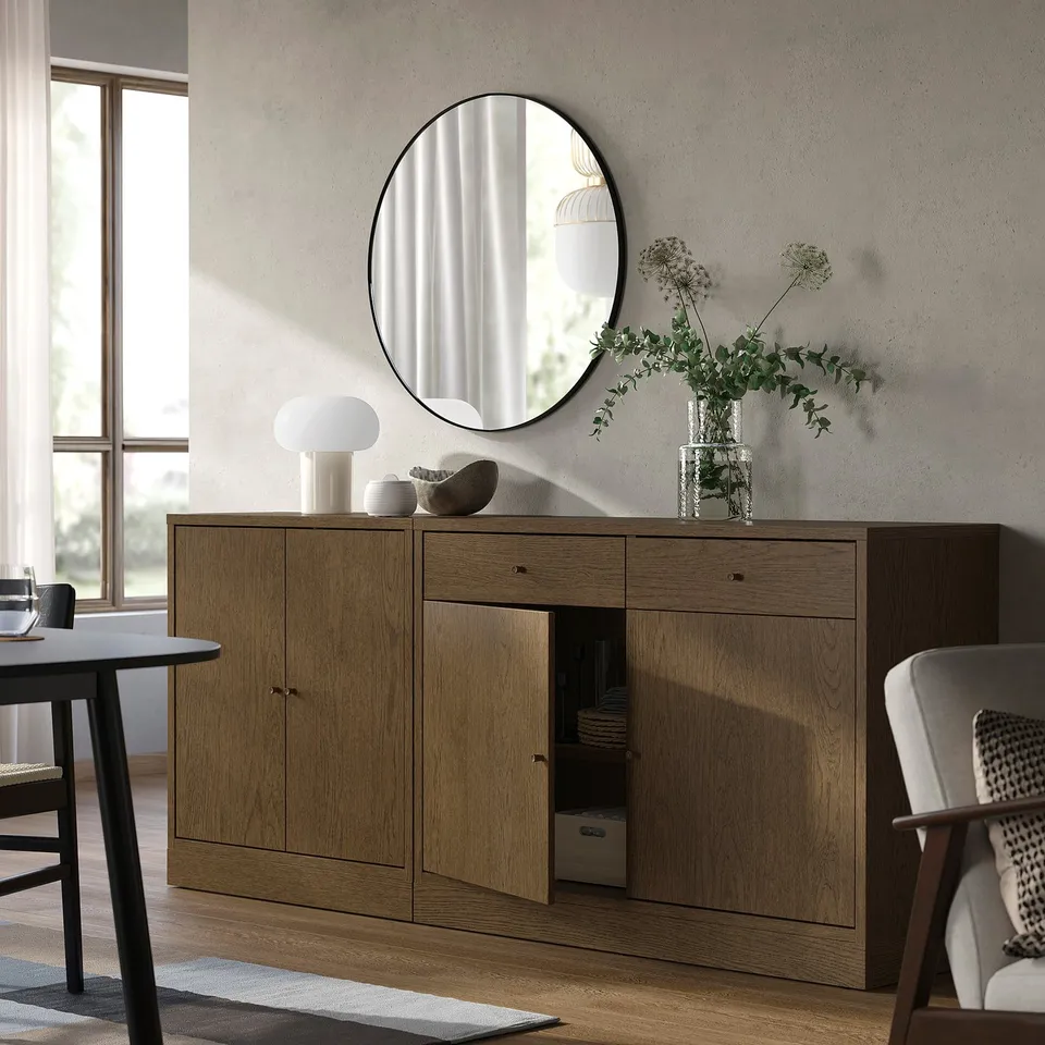Modern dining area with TONSTAD sideboard in wood. Large round mirror above, lamp and vase on top. Open cabinet reveals plates and boxes.
