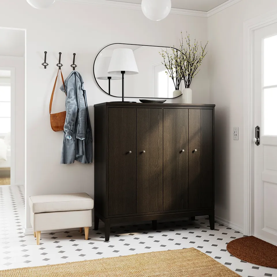 A modern entryway featuring an IDANÄS dark wood cabinet with a mirror, lamp, and hooks. White walls and patterned rug.