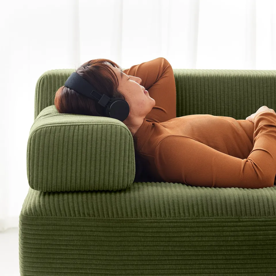 Person relaxes on green JÄTTEBO sofa, listening to headphones, wearing orange outfit.