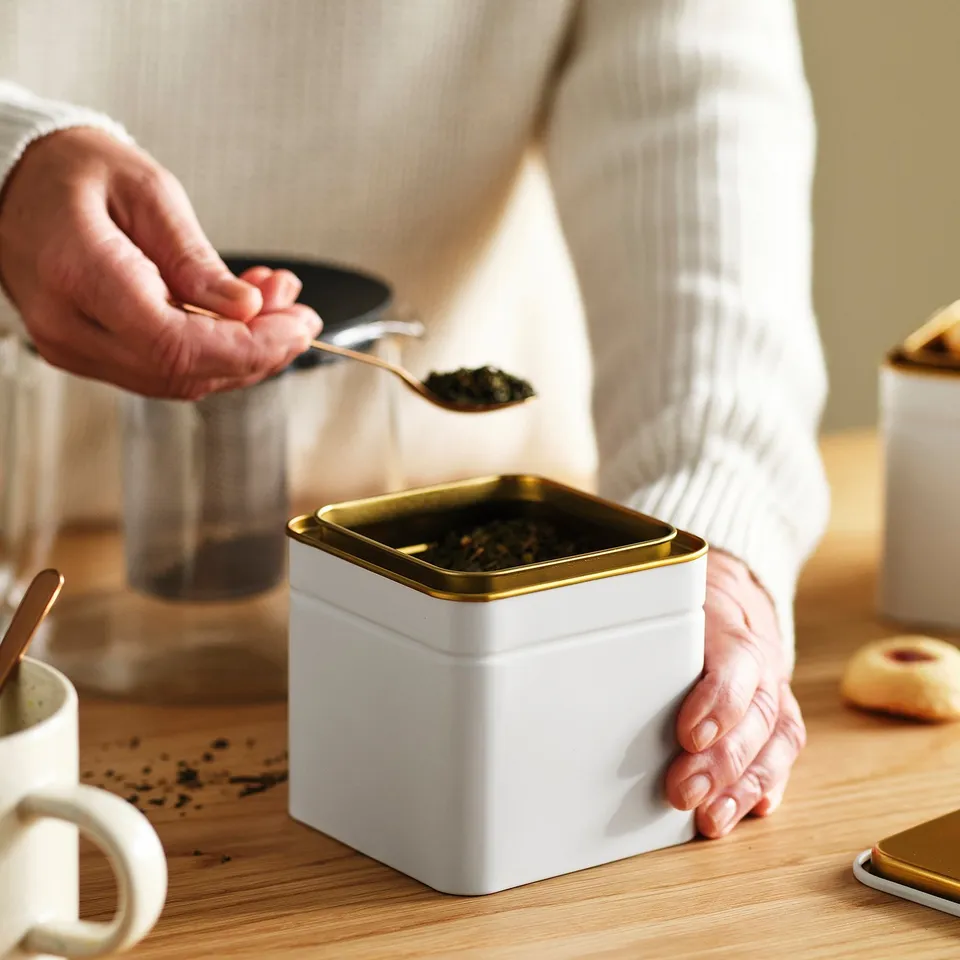 Person scooping tea with gold spoon from white tin can on wooden table.