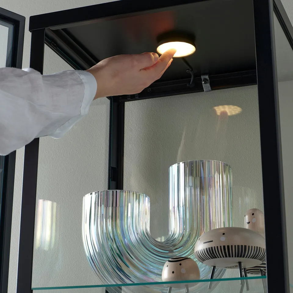 A hand turns on a STRIMSÄV spotlight mounted inside a cabinet, illuminating decorative items on the shelf below.