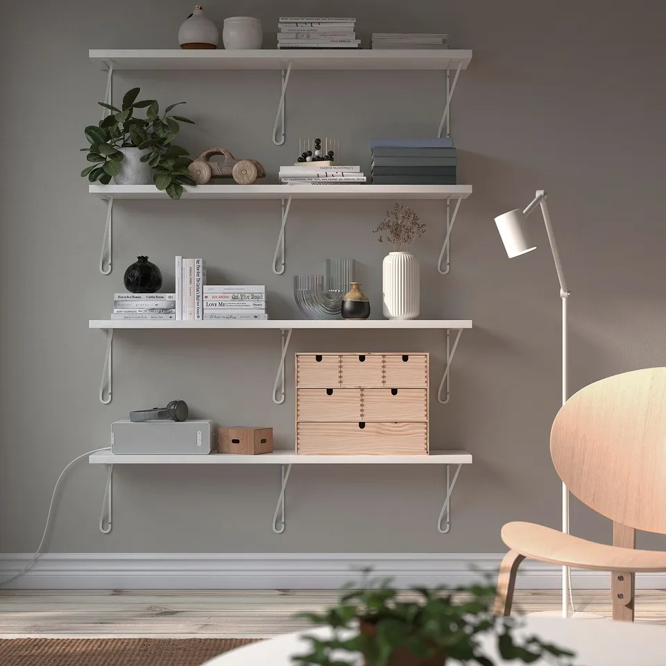 Modern minimalist white BERGSHULT wall shelf, holding decorative items, books, plants, and storage boxes, with white floor lamp and wooden chair.