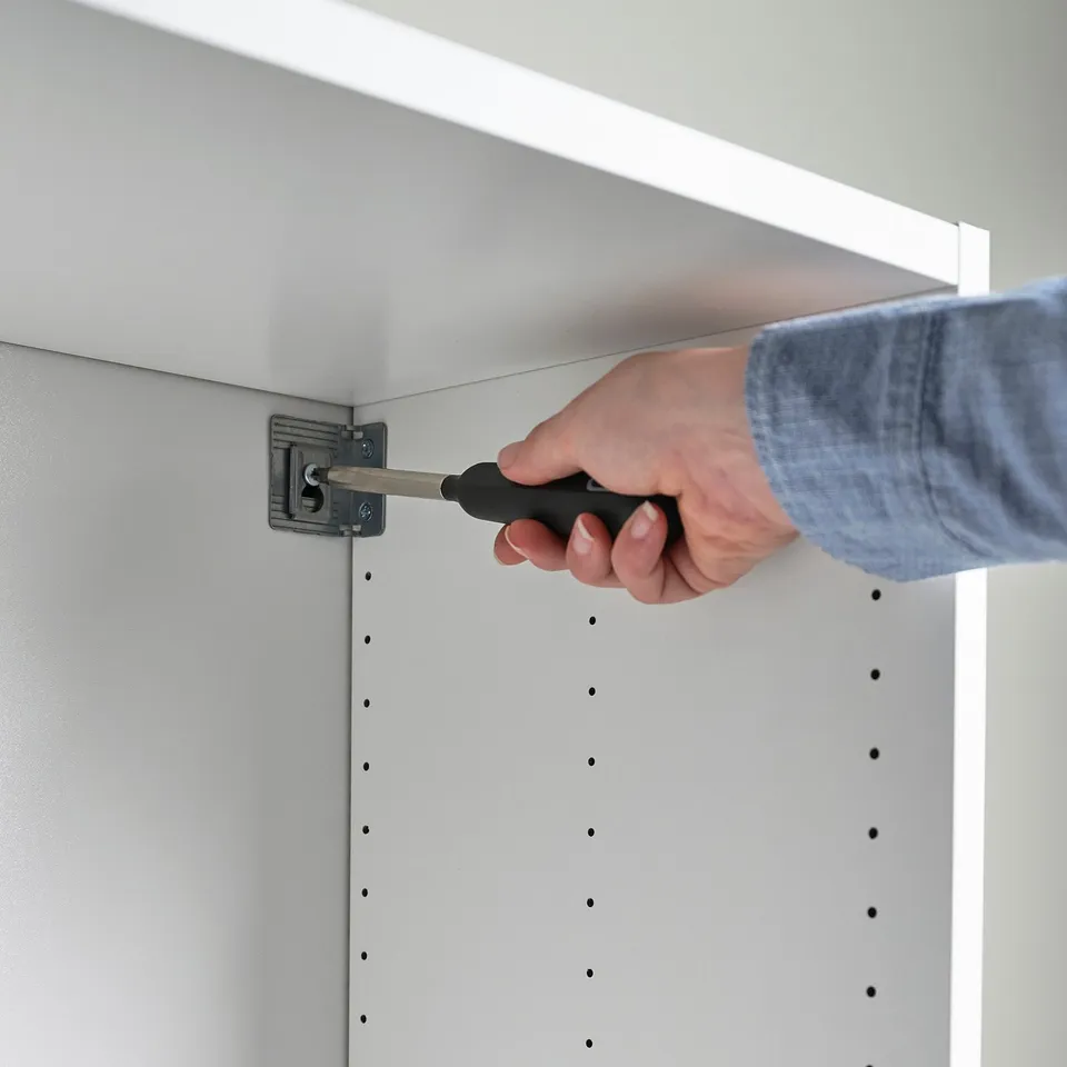 A person is assembling a white cabinet with a screwdriver, securing it to the wall. The cabinet has visible holes for screws.