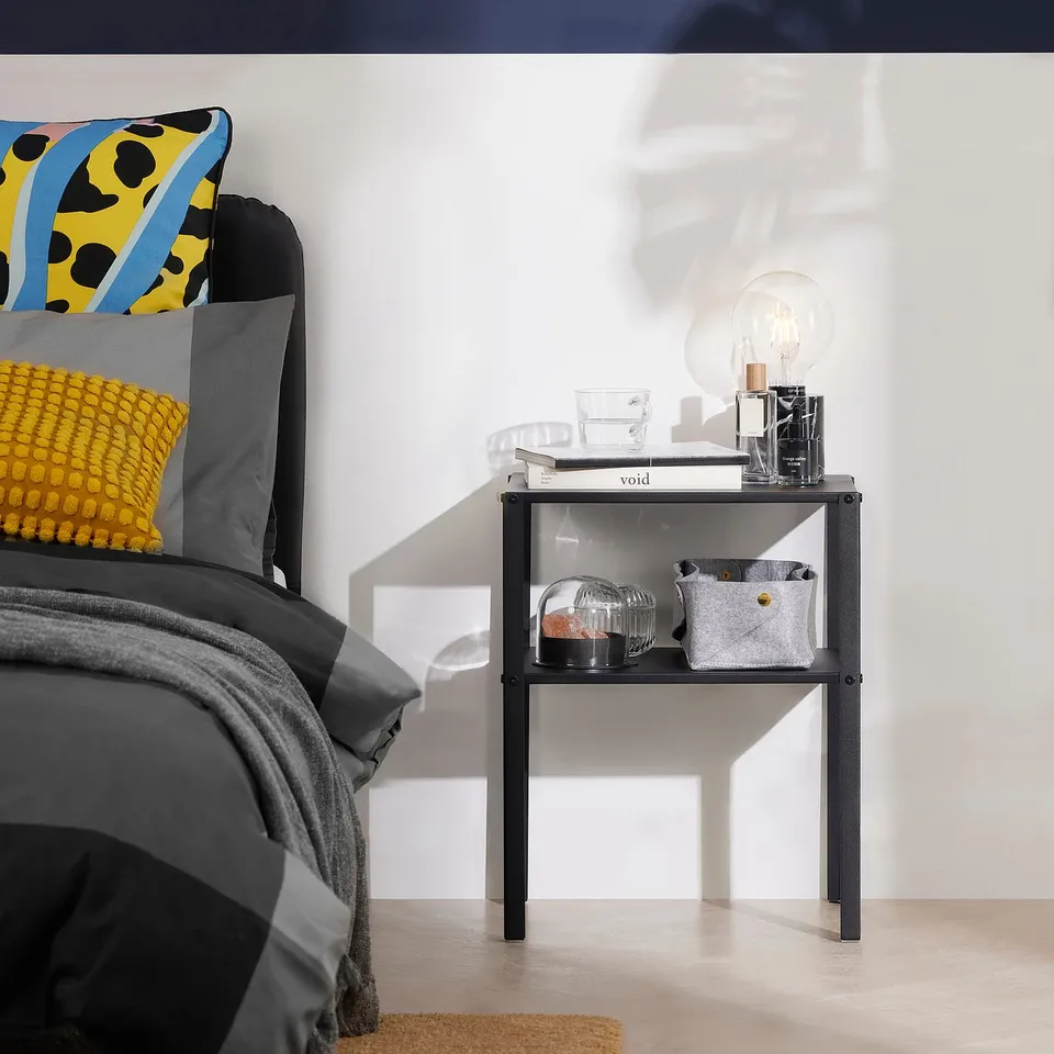 Modern bedroom with KNARREVIK side table holding a light bulb, book, and baskets. Bright pillows decorate the bed.