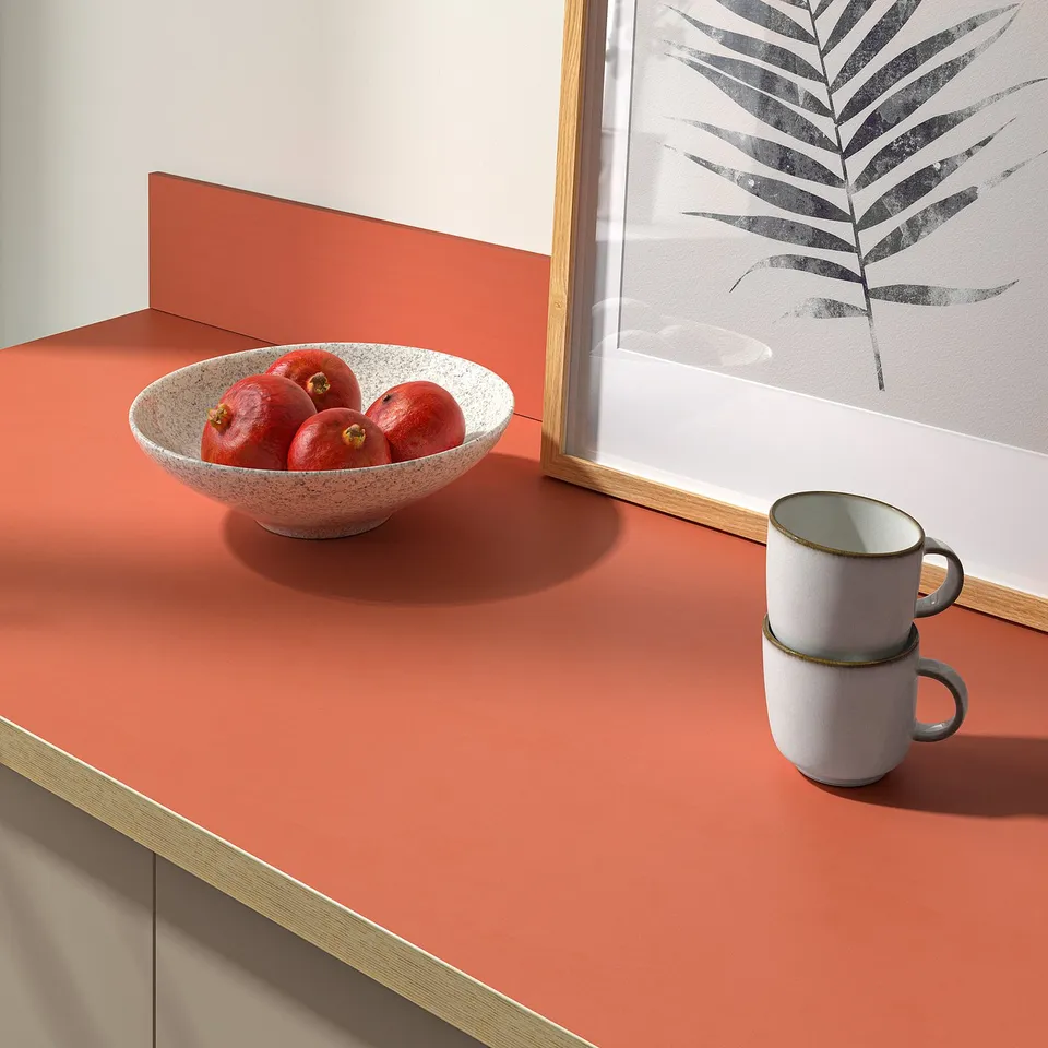 White bowl with red fruits sits on a terracotta-colored countertop, next to stacked white mugs. Beside a framed plant print picture.