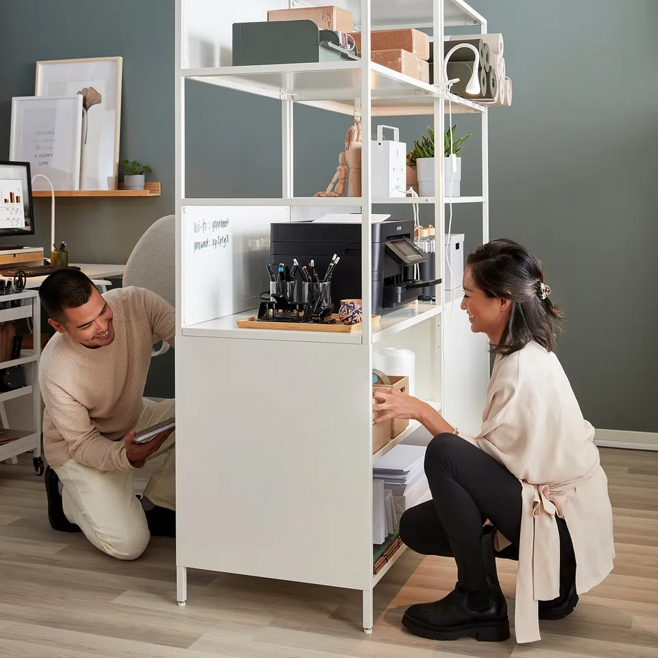 Two people kneeling by large TROTTEN cabinet, room divider with storage, whiteboard, notice board, and office supplies.