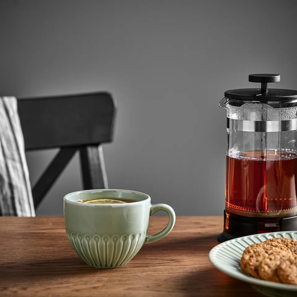 Wooden table with light green mug filled with tea, a cafetière with tea, and a plate with cookies.