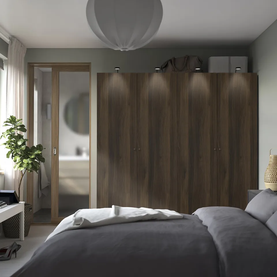 Modern bedroom with large dark wood PAX wardrobe. Mirrored closet door reflects room. Grey bed with white sheets. Potted plant by window.