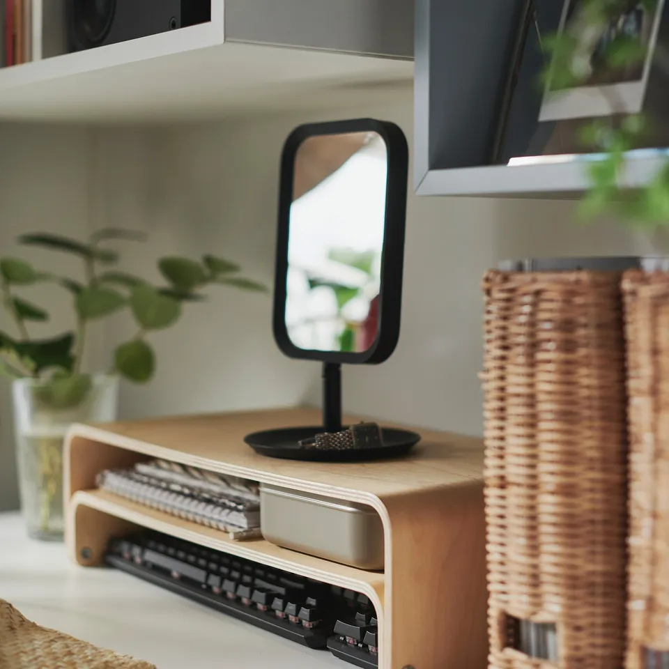 LINDBYN mirror on wooden stand, reflecting greenery, beside radio and wicker basket. Wooden stand holds small items tray.