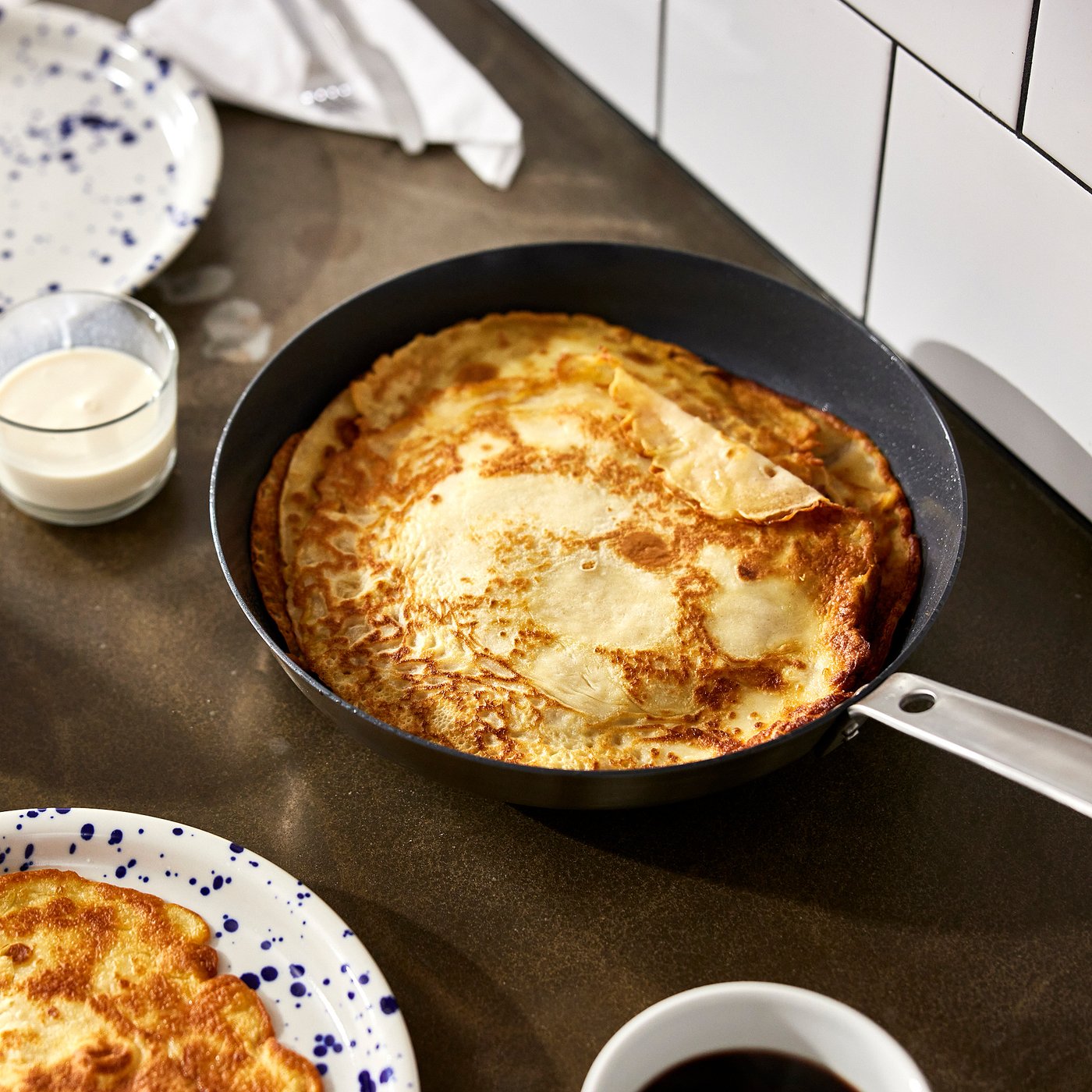 A black frying pan with a golden-brown, puffed pancake sits on a countertop next to plates and cream.