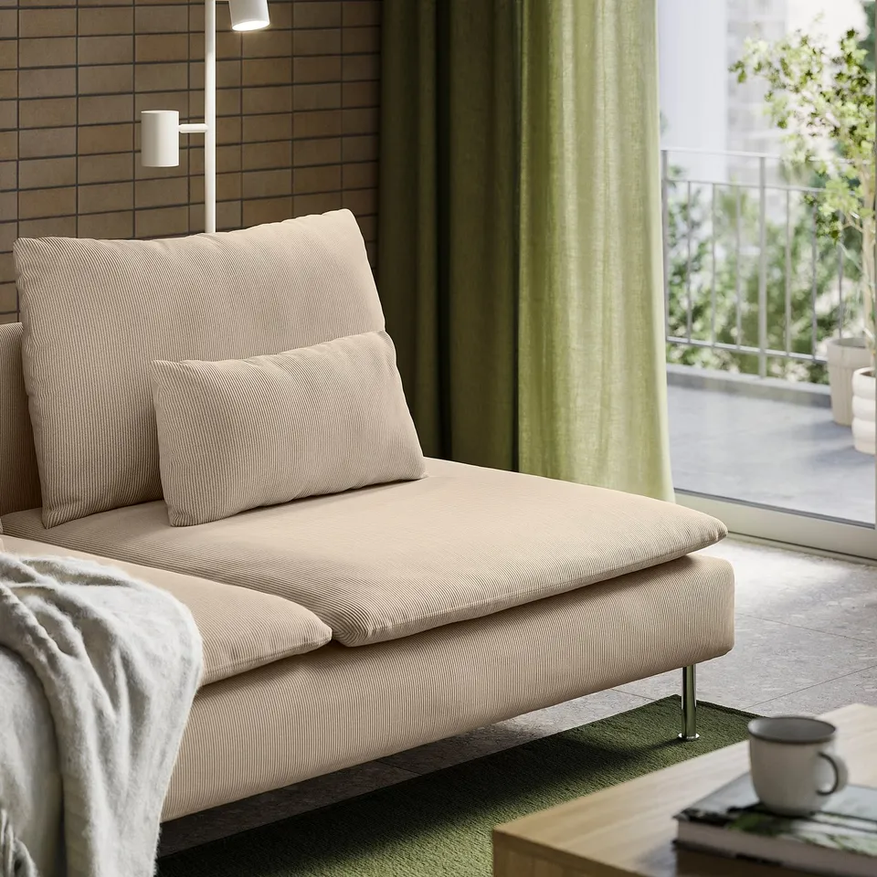 A beige SÖDERHAMN sofa with pillows and a blanket sits against a wooden wall, with a mug and book on a coffee table beside it.