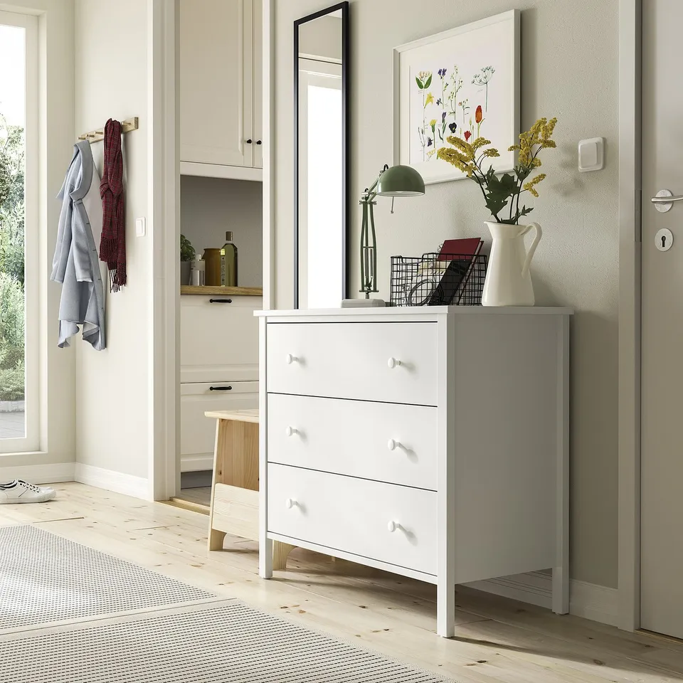 White KILBERGET chest with 3 drawers, lamp, flowers, basket, mirror. Wood floors, grey rug, coats.