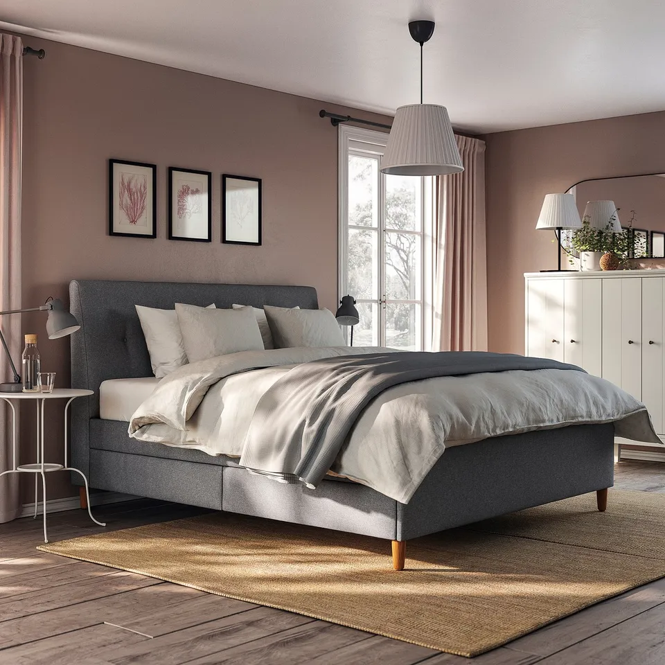 Modern bedroom with IDANÄS grey upholstered bed, white curtains, and pendant light. Neutral decor with rug, plants, and wall art.