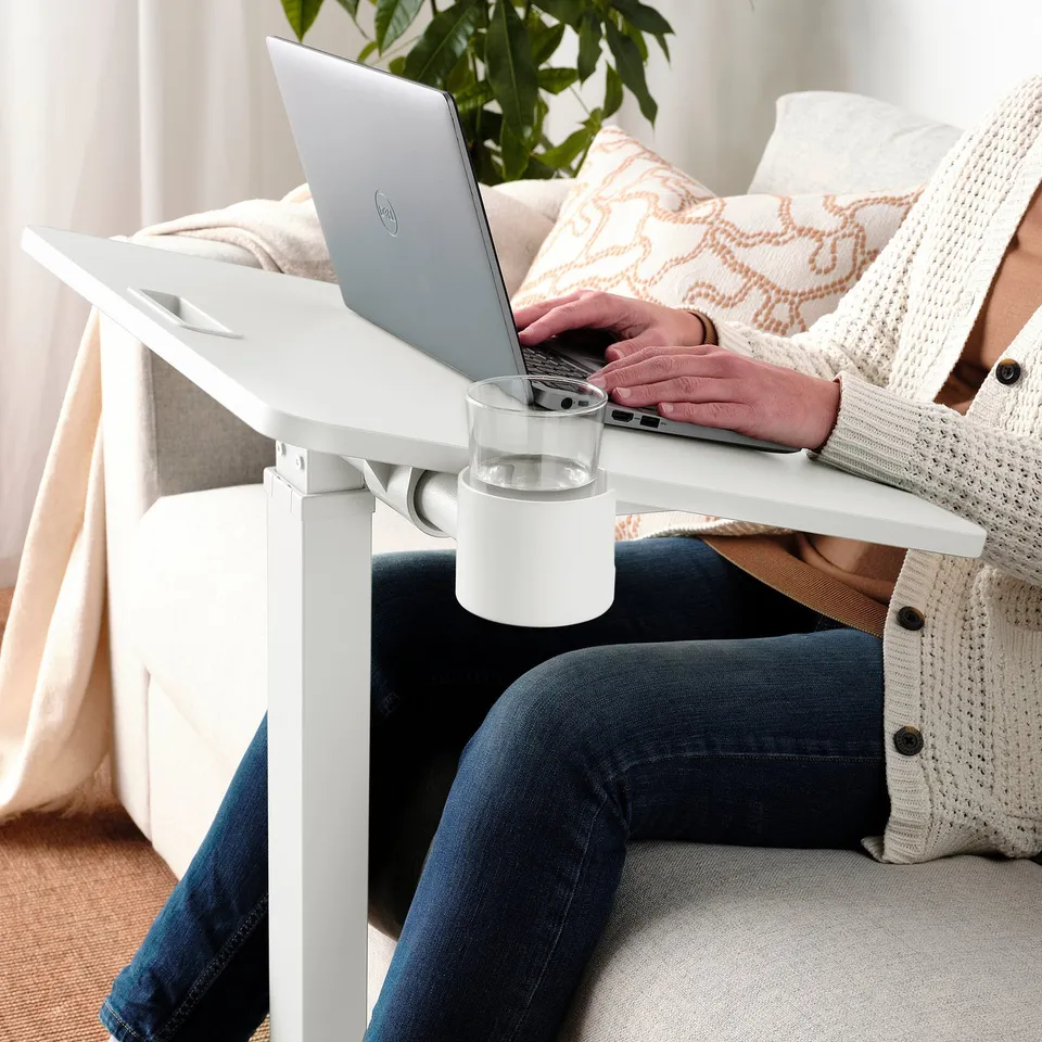 Person working on height-adjustable laptop table with glass holder. Cosy setting with curtain and plant.