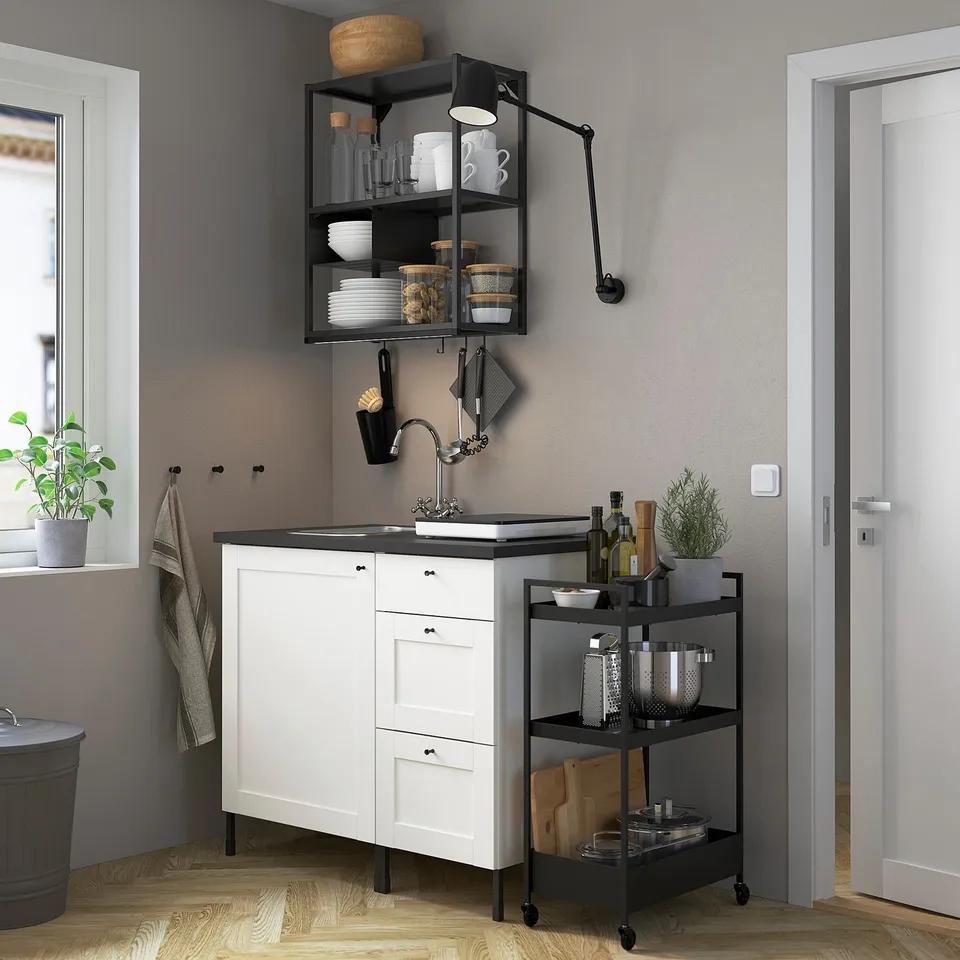 Modern kitchen with white cabinets, black handles, countertop, and metal shelf with jars. Black cart holds kitchen tools and bottles.