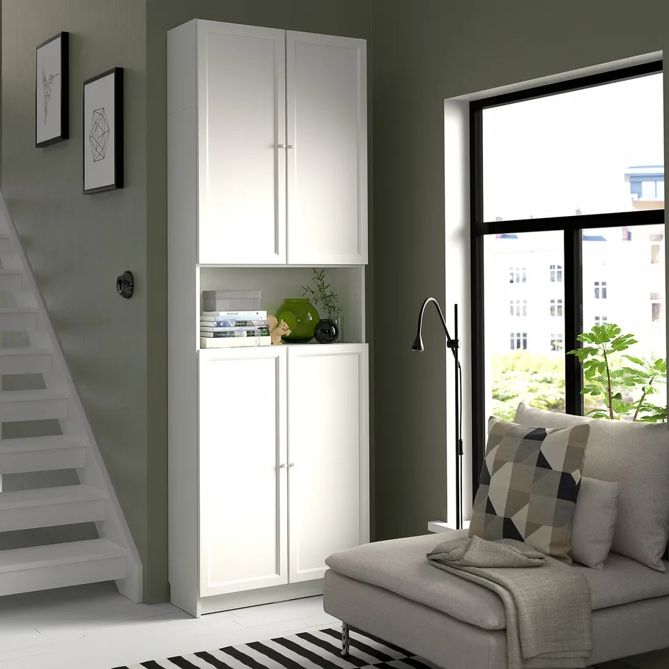 Modern living room with white BILLY bookcase, grey sofa, striped pillows, green wall, large window, stairway, and plant.