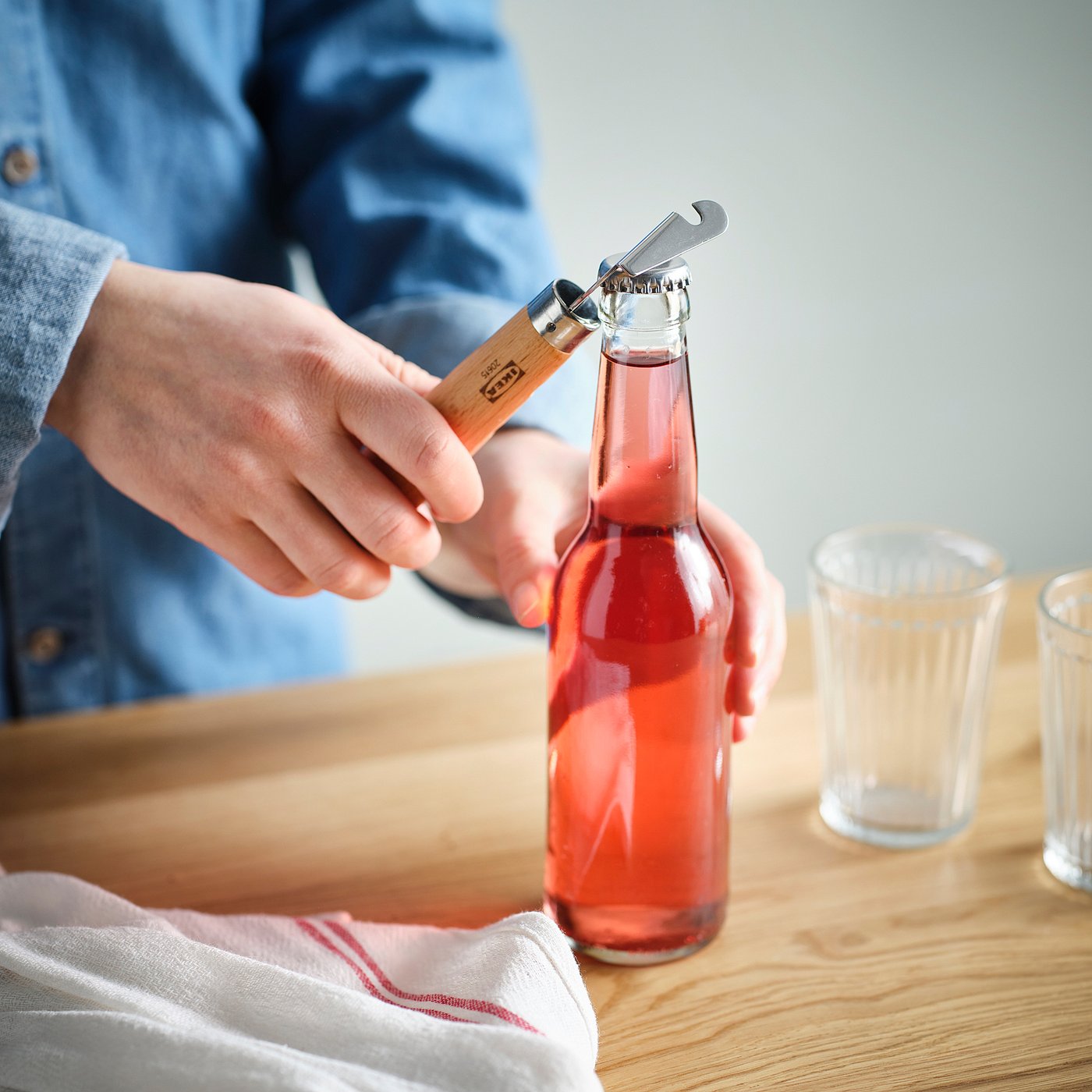 Person opens a red drink bottle using a dual bottle/can opener with a wooden handle on a wooden table.