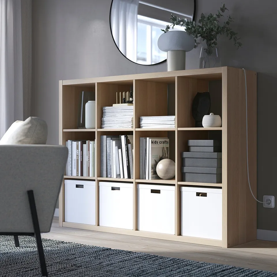 KALLAX shelving unit with books, vases, and storage boxes. Tall, with grey chair, lamp, and plants.