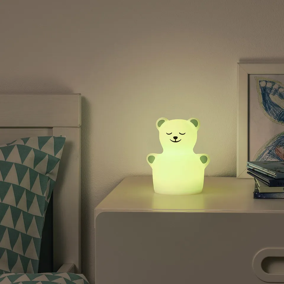 A cosy bear-shaped nightlight on a bedside table, emitting a soft yellow glow, with a patterned pillow and headboard nearby.