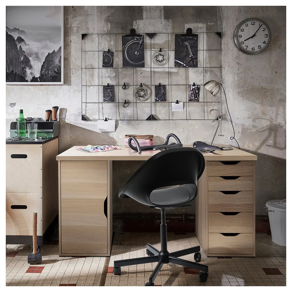 Modern office setup with LAGKAPTEN light brown desk, black swivel chair, wall clock, and decorative wire grid with photos.