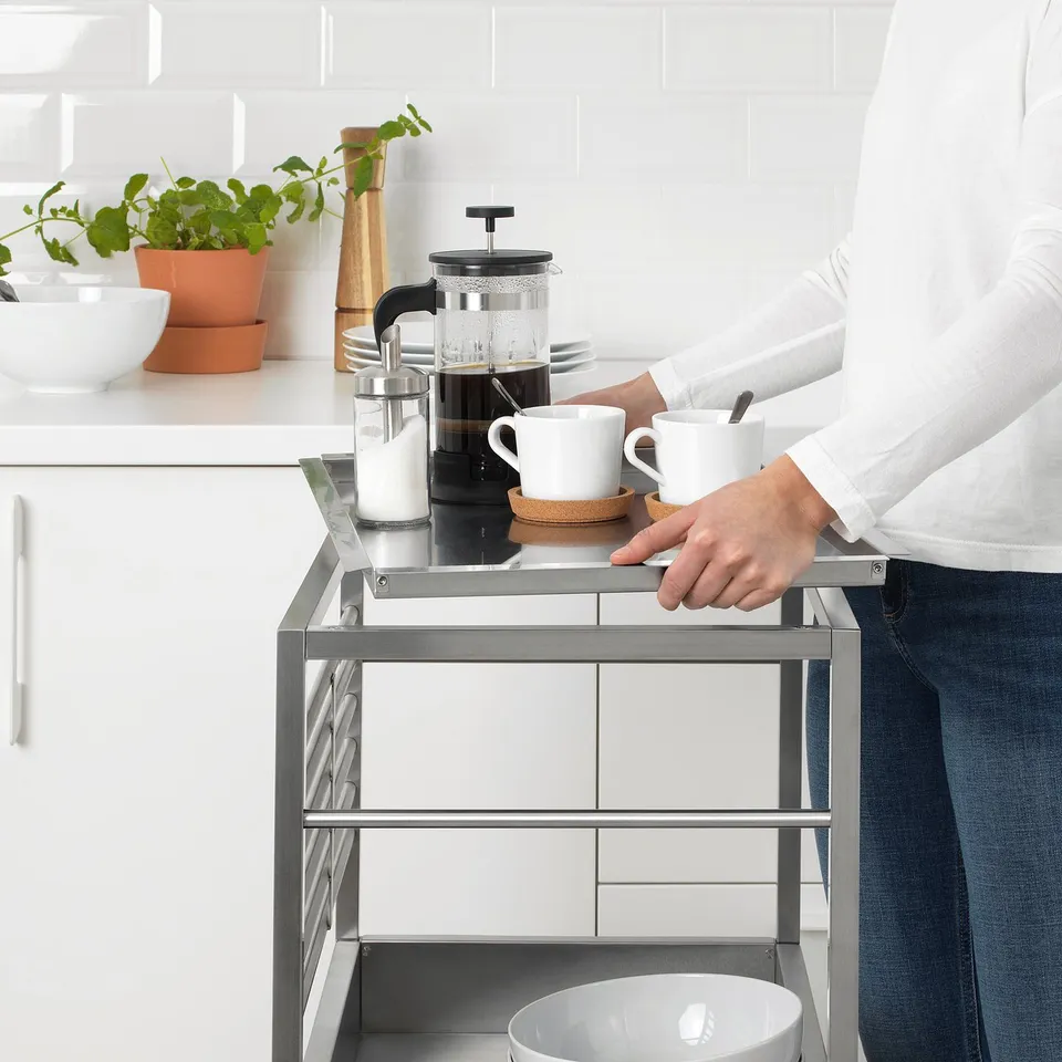 Person pushes metal kitchen trolley with glasses, bowls, and coffee maker on countertop.