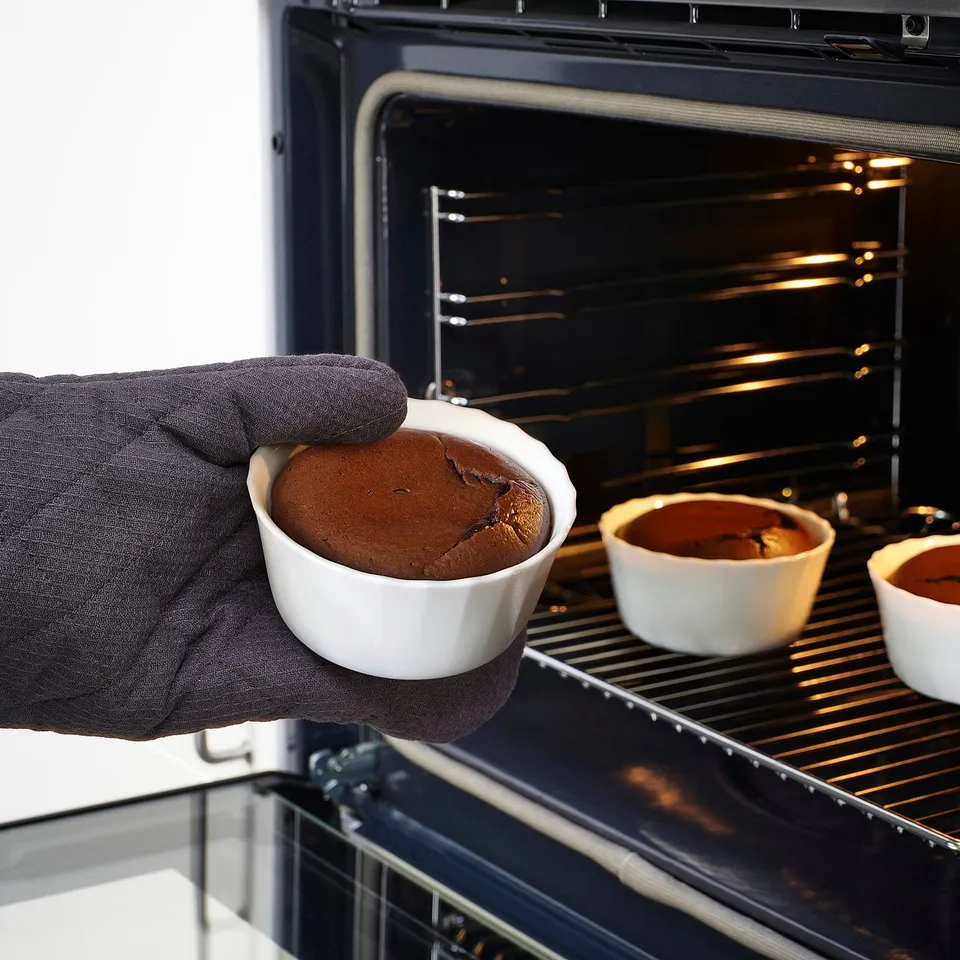 Hand wearing oven mitt holds hot chocolate lava cake in white ceramic dish, being taken out of oven.