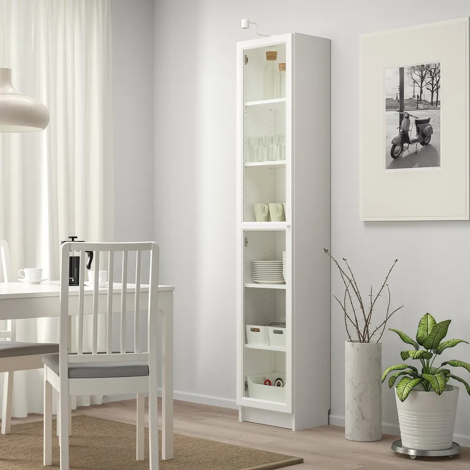 Modern white BILLY glass-door cabinet with cups/dishes in dining area. Black/white scooter, plant, marble vase nearby.