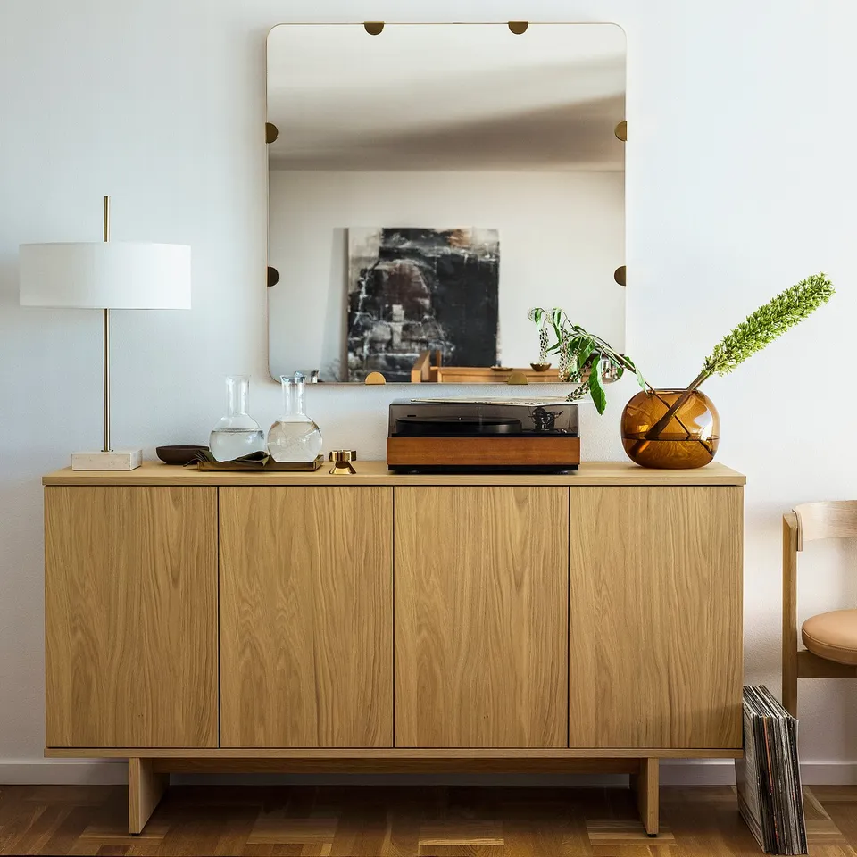 STOCKHOLM 2025 large mirror with brass fittings, oak frame, reflecting room decor. Positioned above a wooden sideboard with turntable, vase, and lamp.