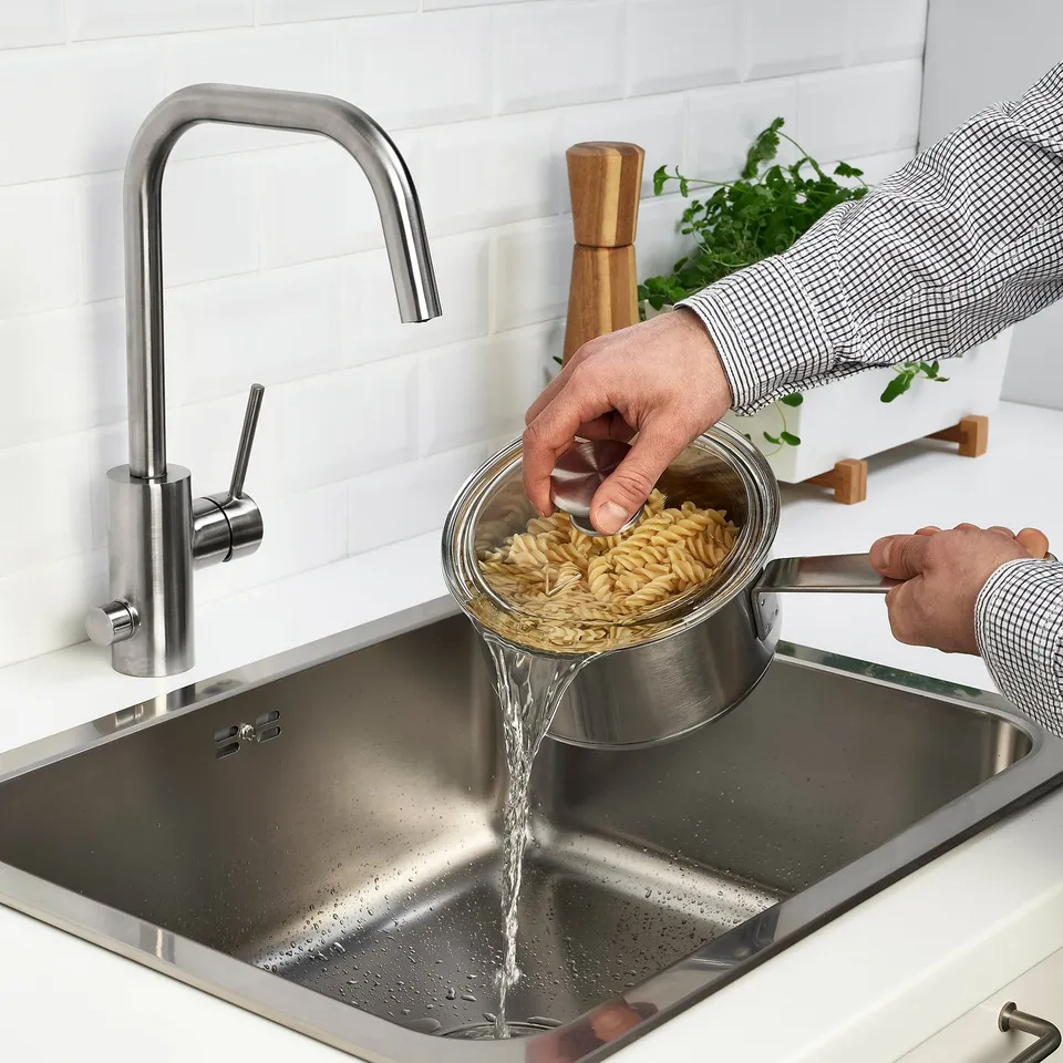Person pours pasta into metal strainer over sink. Modern kitchen setup with stainless tap.