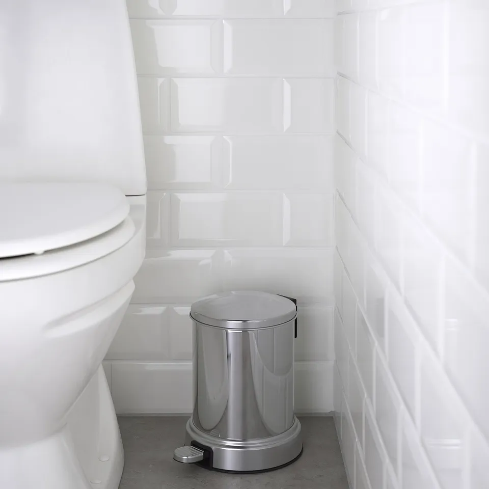 Silver pedal bin beside a white toilet, with a slow-closing lid. The bin has a round shape and chrome details, suitable for bathrooms.