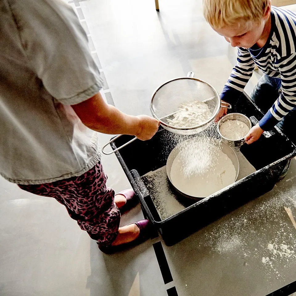Two people prepare food at a counter using bowls and sifters with flour on a black IDEALISK tray.